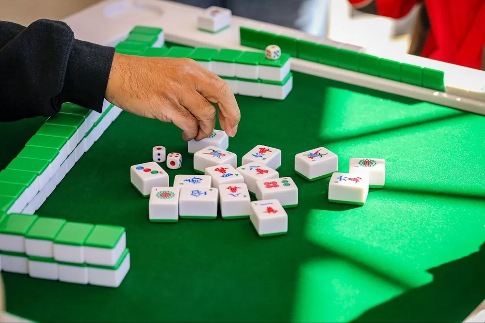A Mahjong set on a felt table with a hand taking a tile.