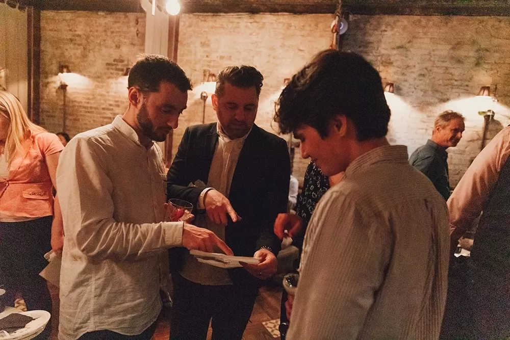 Three men standing and pointing at a piece of paper while they try to solve a puzzle at a dinner party.