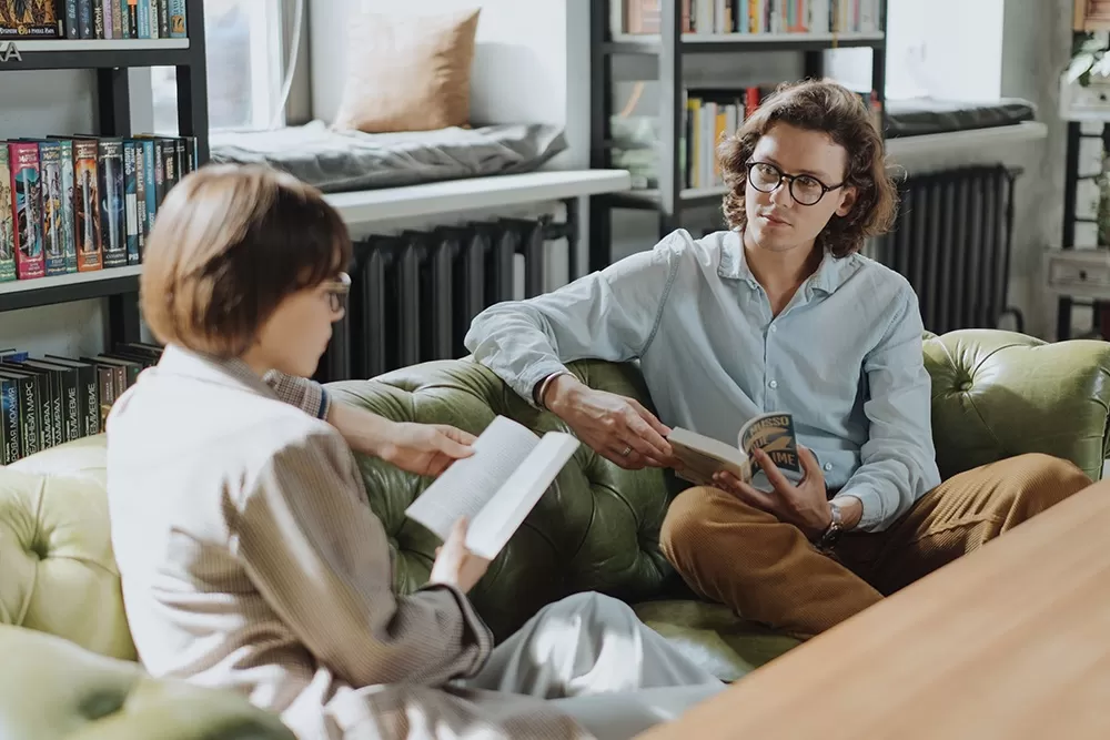 A man with glasses and long hair sitting on a couch looking at a woman read.