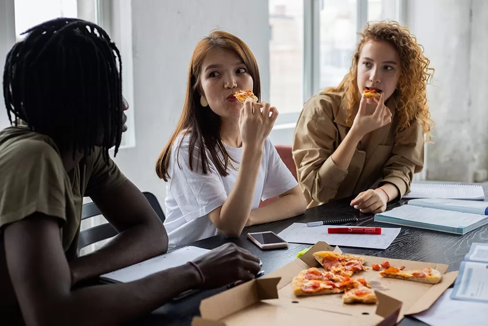 3 women sitting at a desk eating pizza looking like they are interested in what the others are saying.