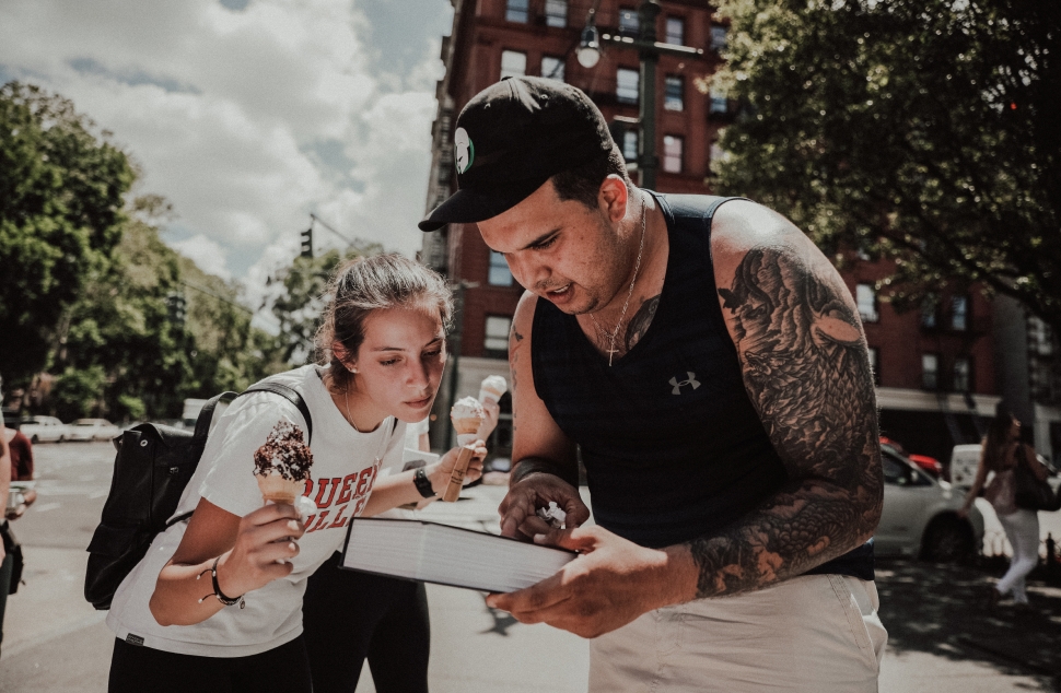 A heavily tattooed man helping a woman holding ice cream cones solve a puzzle in the streets of New York City.