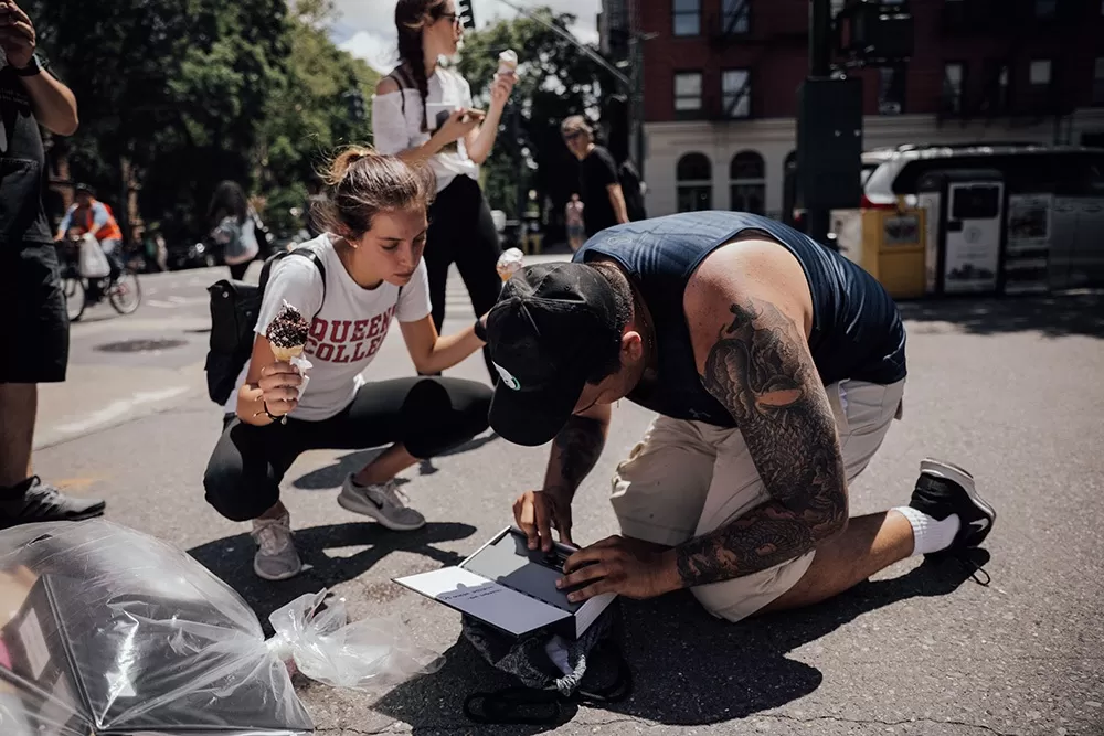 A woman squatting while holding ice cream cones looking at a man that is kneeling on the ground trying to solve a puzzle box.